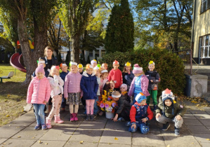 Dzieci z grupy Żuczków, w opaskach z kolorowymi jesiennymi listkami pozują do zdjęcia grupowego z bukietem jesiennych kwiatów, w ogrodzie przedszkolnym.
