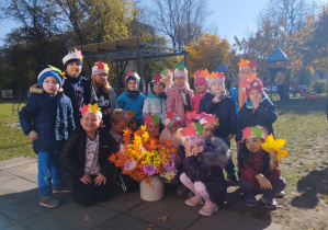 Dzieci z grupy Żabek, w opaskach z kolorowymi jesiennymi listkami pozują do zdjęcia grupowego z bukietem jesiennych kwiatów, w ogrodzie przedszkolnym.