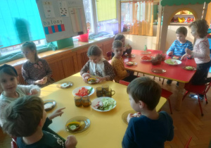 Dzieci z grupy Żuczków sieddzą przy żółtym i czerwony stole, sięgają ulubione warzywa, którymi ozdabiają zimową kanapkę.