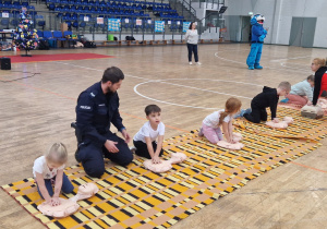 Dzieci na siedząna kocu w pozycji klęcznej, przygotowują się do zaprezentowania prawidłowego sposobu wykonywania masażu serca i sztucznego oddychania.