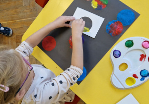 Dziewczynka siedzi przy żółtym stole i stempluje szablon kropki przy użyciu kolorowych farb i pomponów kreatywnych.