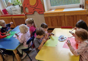 Dzieci malują brązową farbą rolki po papierze, tworząc pień dzewa.