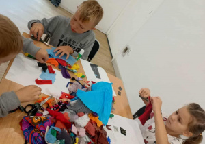 Dzieci siedzą przy stole i wycinają materiałowe elementy do portretu