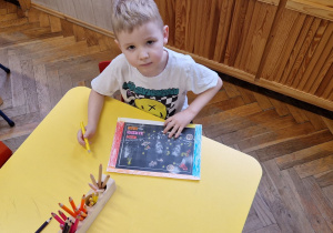 Chłopiec siedzi przy żółtym stole i przygotowuje, koloruje list.