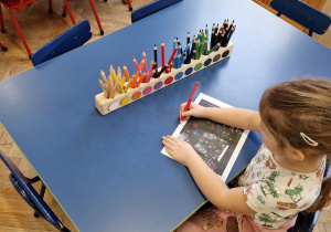 Dziewczynka siedzi przy niebieskim stole i przygotowuje, koloruje list.