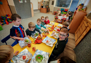 Chłopcy i dziewczynki siedzą przy żółtym stole i ozdabiają swoje kanaki warzywami ułożonymi na półmiskach.