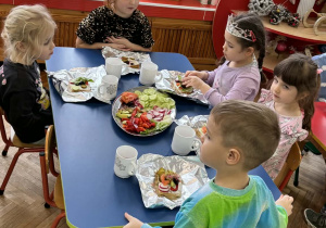 Chłopcy i dziewczynki siedzą przy niebieskim stole i ozdabiają swoje kanaki warzywami ułożonymi na półmiskach.