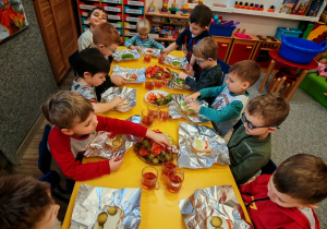 Chłopcy i dziewczynki siedzą przy żółtym stole i ozdabiają swoje kanaki warzywami ułożonymi na półmiskach.