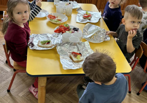 Chłopcy i dziewczynki siedzą przy żółtym stole i ozdabiają swoje kanaki warzywami ułożonymi na półmiskach.