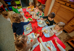 Chłopcy i dziewczynki siedzą przy czerwonym stole i ozdabiają swoje kanaki warzywami ułożonymi na półmiskach.
