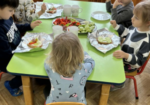 Chłopcy i dziewczynki siedzą przy zielonym stole i ozdabiają swoje kanaki warzywami ułożonymi na półmiskach.