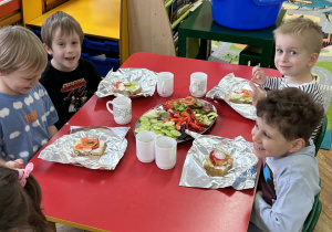 Chłopcy i dziewczynki siedzą przy czerwonym stole i ozdabiają swoje kanaki warzywami ułożonymi na półmiskach.