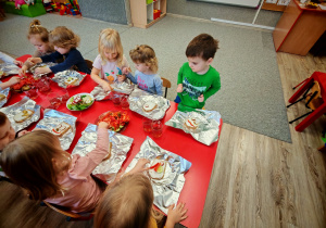 Chłopcy i dziewczynki siedzą przyczerwonym stole i ozdabiają swoje kanaki warzywami ułożonymi na półmiskach.