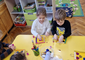 Dzieci siedzą przy żółtym stole i ozdabiają ramki.