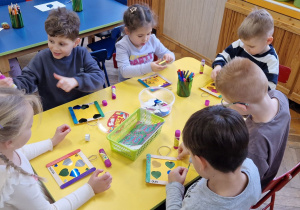 Dzieci siedzą przy żółtym stole i ozdabiają ramki.