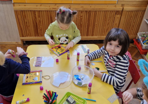 Dzieci siedzą przy żółtym stole i ozdabiają ramki.